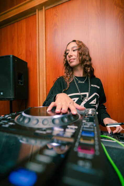 A person using a DJ controller in front of a wooden wall, with one hand on the controls and a speaker in the background.