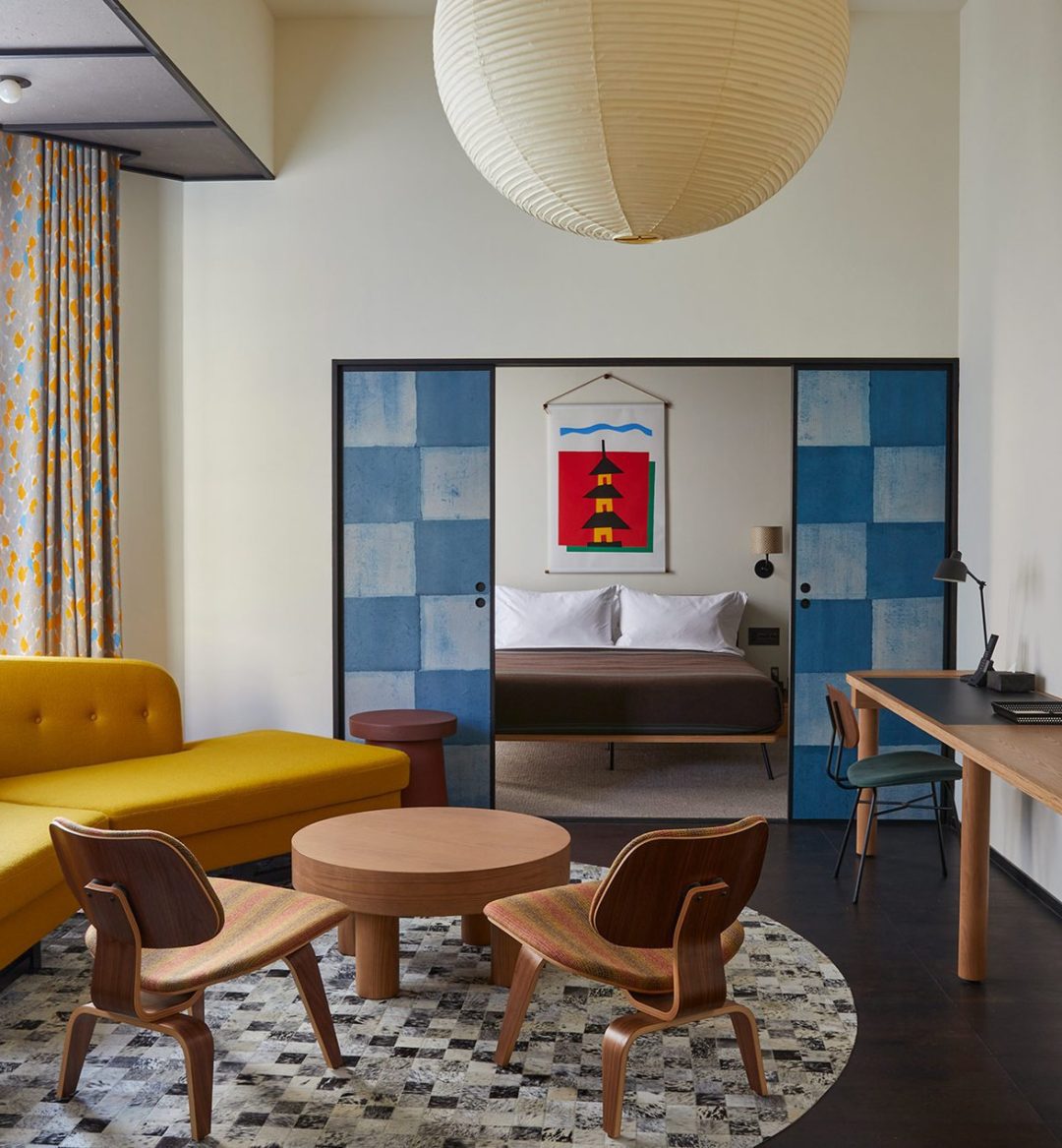 A large living space with a stylish yellow sofa and large traditional light fixture opens onto a dedicated bedroom