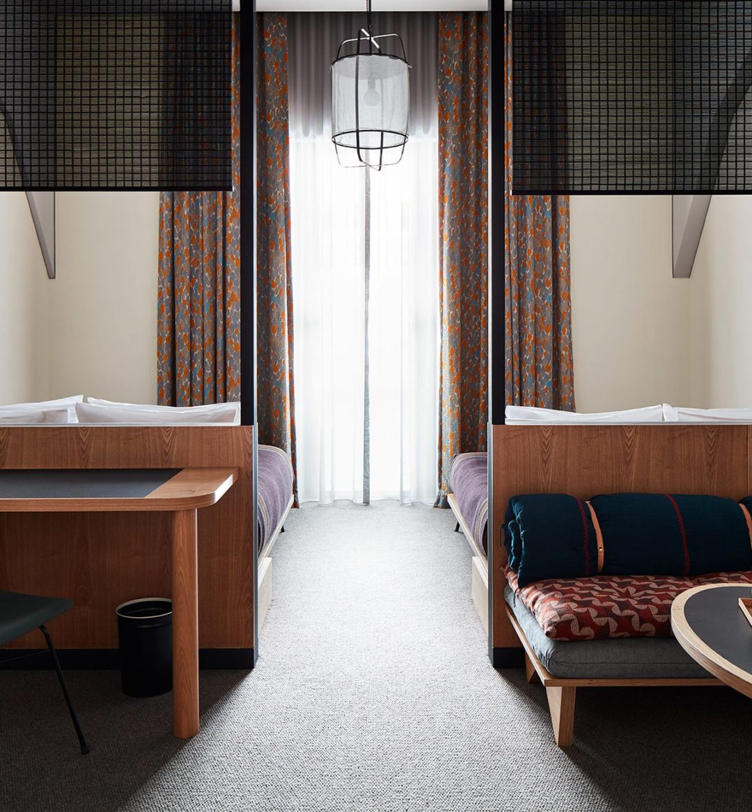 Two beds are separated by a space with a traditional light fixture suspended from the ceiling; a desk on one side of the room provides space for work