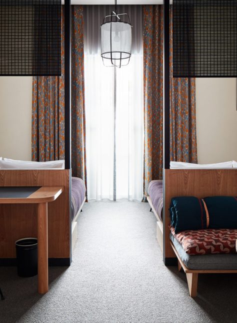 Two beds are separated by a space with a traditional light fixture suspended from the ceiling; a desk on one side of the room provides space for work