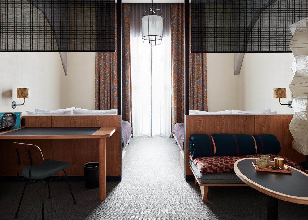Two beds are separated by a space with a traditional light fixture suspended from the ceiling; a desk on one side of the room provides space for work