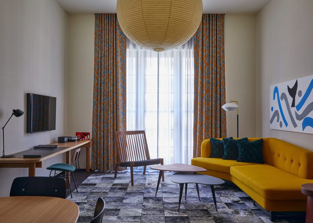 A large living space with a stylish yellow sofa and large traditional light fixture features a window providing natural light to the entire room