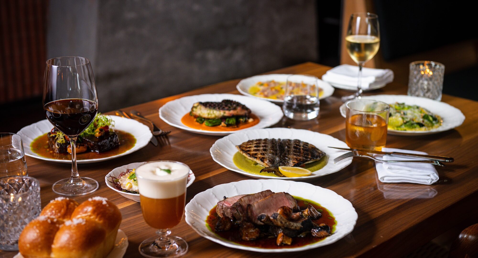 A table set with plates of gourmet dishes, wine glasses, a cocktail, and bread rolls in a restaurant setting.