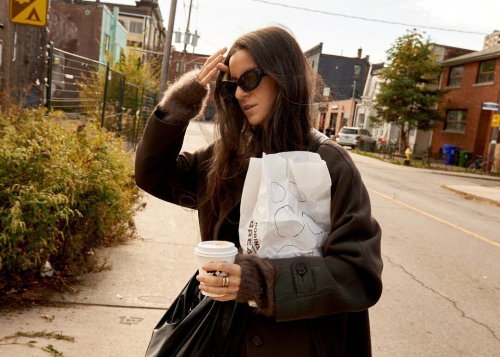 Woman in sunglasses walks down a city street holding a coffee cup and a paper bag, with brick buildings in the background.